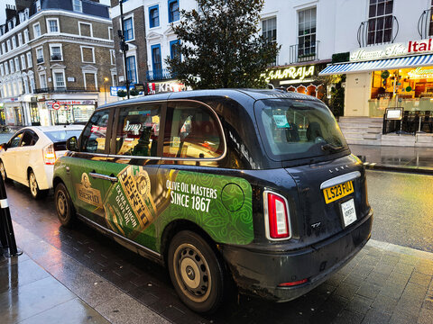 Black London LEVC taxi with Filippo Berio olive oil advertising on a rainy city street in London, UK