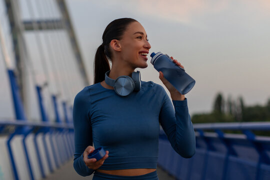 Happy young caucasian woman drinking water after exercise on modern pedestrian bridge