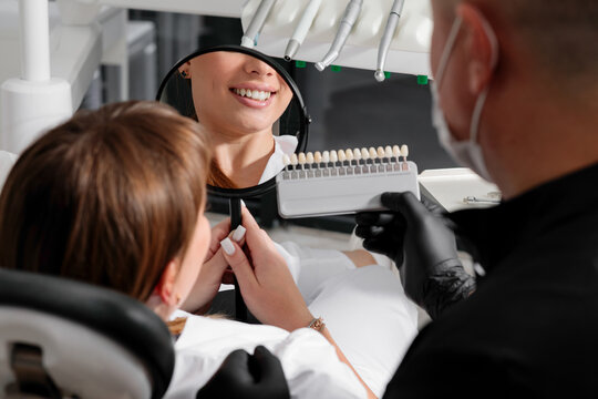 a smiling woman at a dental clinic holds a mirror while a dentist in black gloves shows her a tooth shade guide