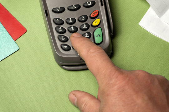 A hand is pressing the number nine on a payment terminal. The terminal is on a green surface and cards are nearby. The setting looks like a retail store during a weekday