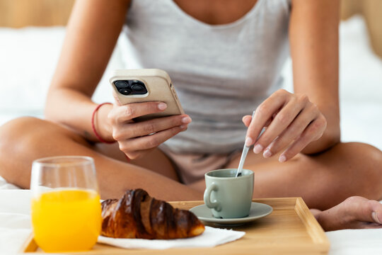 Woman using a smartphone while having breakfast on bed at home.
