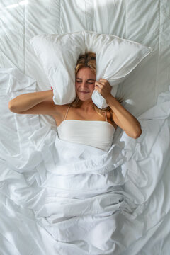 Tired woman covering her ears with a pillow in bed at home.