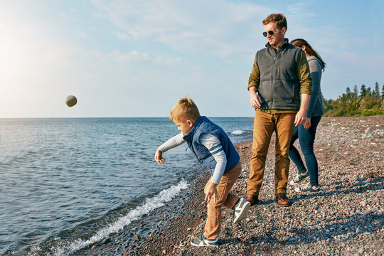 Parents, child and rock throwing into lake with bonding, vacation and travel together. Man, woman and boy kid tossing stone into water for fun, playful games and lakeside family holiday outdoor