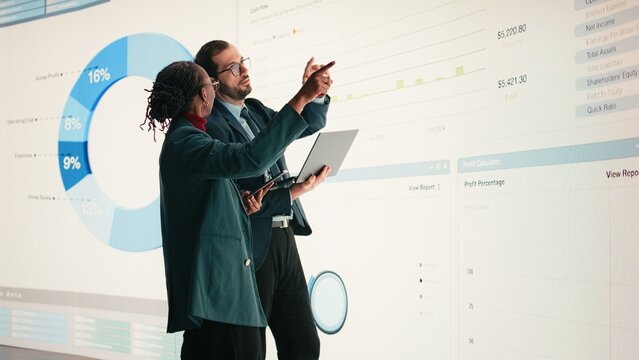 Corporate managers evaluate fiscal performance metrics on digital wall, collaborating, Diverse executive team discusses quarterly earnings report and shareholder equity charts during boardroom.