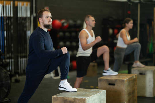 Focused young bearded man in comfortable sportswear participating functional cross training at gym, stepping up onto wooden box to work glutes, quads and hamstrings ..