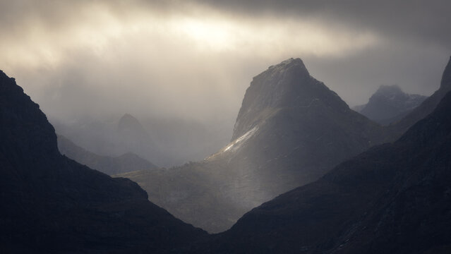 View of Lofoten mountains with dramatic peaks illuminated by a shaft of sunlight breaking through heavy clouds and mist in Vagan Municipality, Nordland, Norway.