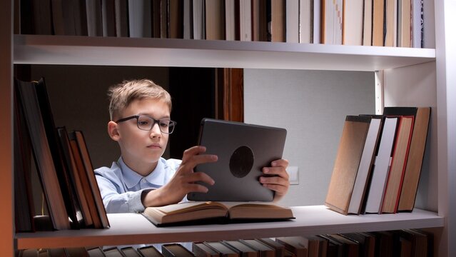 Young boy in glasses focusing on a digital tablet screen, sitting at a bookshelf with traditional books, representing modern technology complementing education