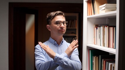 Man in light blue shirt and glasses stands in a library with arms crossed in an emphatic stop gesture, serious expression conveying refusal, disagreement and boundary setting © Aliaksandr