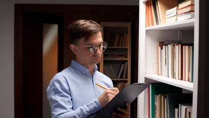 Man wearing glasses intently focusing, writing notes with a pencil on a clipboard while standing by a bookshelf filled with books in a library, conducting inventory or research © Aliaksandr