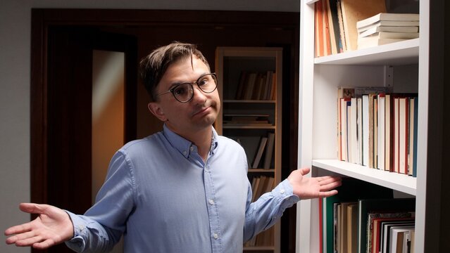 Young man in glasses shrugging with open hands, showing a puzzled or uncertain expression while standing beside a bookshelf filled with books in an indoor setting