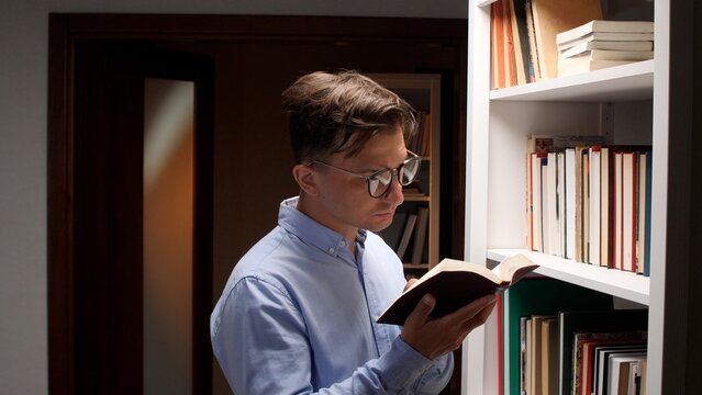 Man with glasses standing in a home library, focused on reading a book beside a bookshelf filled with titles, quietly studying and enjoying lifelong learning and research