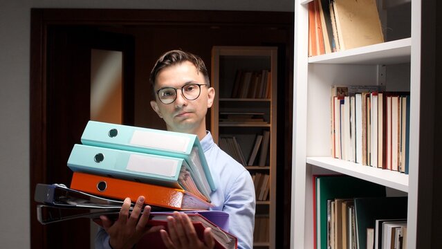 Overwhelmed businessman with glasses carrying a precarious pile of office binders and documents, showing bureaucracy, paperwork, and workload demands in an office environment