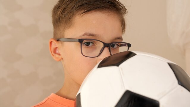 Young boy wearing dark framed glasses holding a black and white soccer ball, looking directly at the camera with a serious expression, showcasing childhood, sport, and hobby indoors