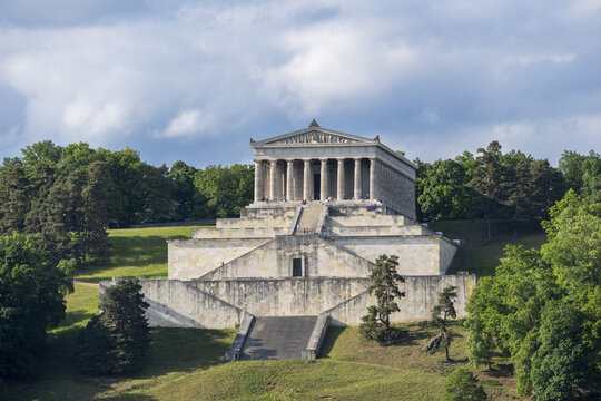 View of the Walhalla memorial temple with people on the monumental stone staircases and neoclassical columns surrounded by lush green forest Donaustauf, Bayern, Germany.