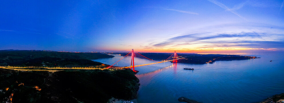 Panoramic aerial view of illuminated Yavuz Sultan Selim Bridge at night in Istanbul, Turkey. Red lights on modern suspension bridge over Bosphorus connecting Europe and Asia at dusk twilight.