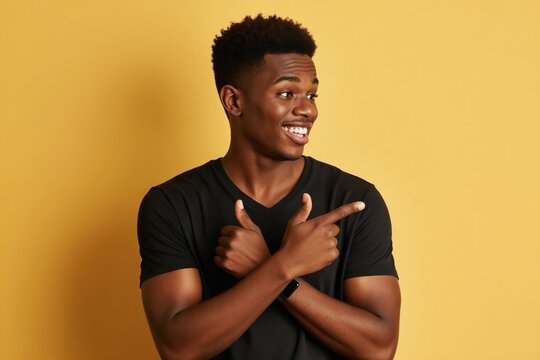 Young black man with smart watch points fingers to side with happy smile. Male model gestures and looks away, wearing black t-shirt on yellow background. Copy space available.