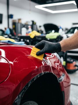 Professional technician's hand in black glove applying paint protection film to a red car fender in a clean auto detailing workshop, showcasing vehicle maintenance and protection.
