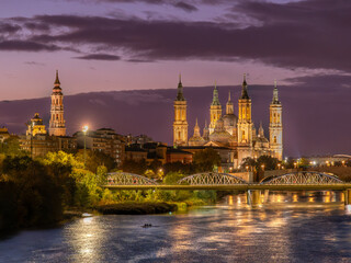 Fototapeta premium Blue hour tourism view of the Basilica of Our Lady of the Pillar in Zaragoza, Spain, illuminated above the Ebro riverfront and nearby urban gardens.