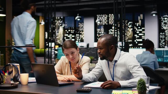 Corporate staff collaborating in a shared workspace late at night, engaged in teamwork and planning sessions. Multiethnic group uses laptop to analyze data and discuss strategies, action plan.