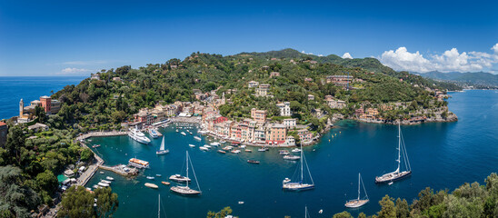 Fototapeta premium Panoramic view of beautiful Portofino on a springtime afternoon with harbor, boats, yachts, waterfront, villas, green hills, and clear blue water on the Ligurian coast in Italy against blue sky