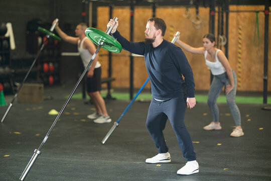 Focused young bearded man in comfortable sportswear performing single arm landmine press during group cross training in well-equipped gym, aiming to strengthen body muscles