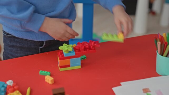 Boy student building colorful plastic blocks with hands on a red table in a preschool classroom surrounded by crayons and drawings; concentration creativity.