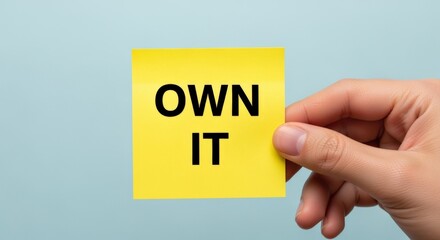Obraz premium Hand Holding Yellow Sticky Note with 'Own It' Text Against Blue Background