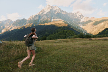 Naklejka premium Woman hiking through a grassy meadow toward rugged mountains, carrying a backpack, enjoying fresh air and wide landscapes in a nature adventure scene.
