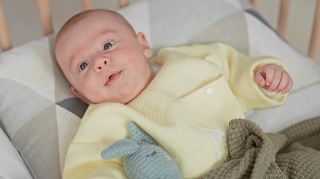 Baby in yellow sleeper lies in a wooden crib looking up with hand raised in a building; gentle curiosity.