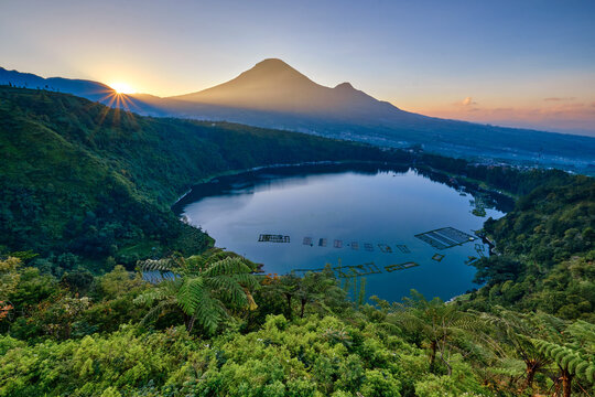 View of Love Valley Seroja Hill overlooking Telaga Menjer lake with fish cages and Mount Sindoro under a colorful sky in Garung, Wonosobo, Central Java, Indonesia.