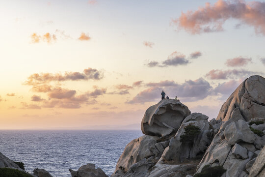 View of people standing on the granite rock formations of Capo Testa overlooking the sea under a sunset sky with pink clouds in Capo Testa, Sardegna, Italy.