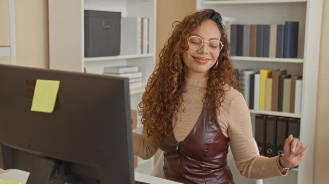 Woman smiling and raising hands at a desk computer in office with bookshelves, binders and sticky note on monitor; celebration success.