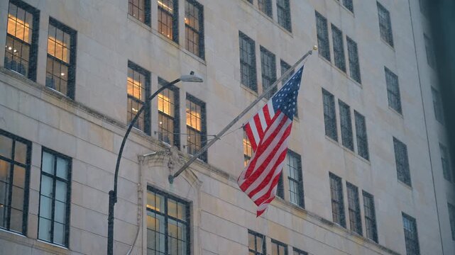 American Flag on Classical Manhattan Building During Snowstorm. American flag on flagpole mounted on beige stone building facade, snowfall, lit windows, street lamp, low angle view