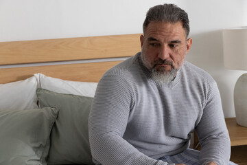 Fototapeta premium Mature man sitting on bed edge, looking right, wearing sweater and plaid pajama pants near lamp