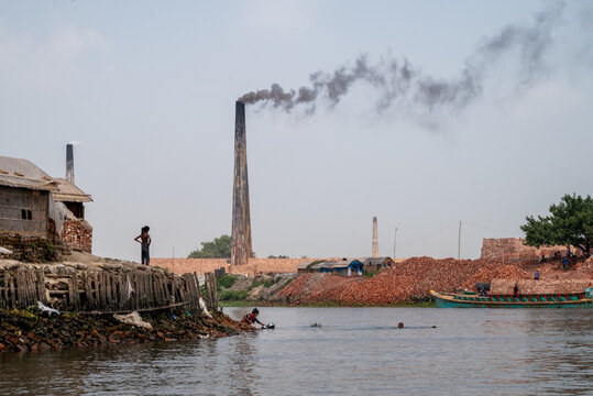 Narayanganj, Bangladesh - 12 April 2026: View of brick kilns with tall chimneys emitting smoke along the riverbank with piles of red bricks and people.