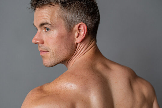 Bare male shoulder and upper back turned from soft studio light, warm skin highlight and shadow with visible neck contours and textured skin expression of calm confidence
