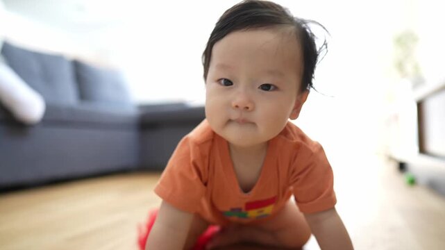 Soft light fills a cozy living room. Baby sits on wood floor, wearing orange shirt. Red tulle drapes around baby's legs. Baby smiles, looks up with curious eyes. Calm, joyful, innocent moment captured