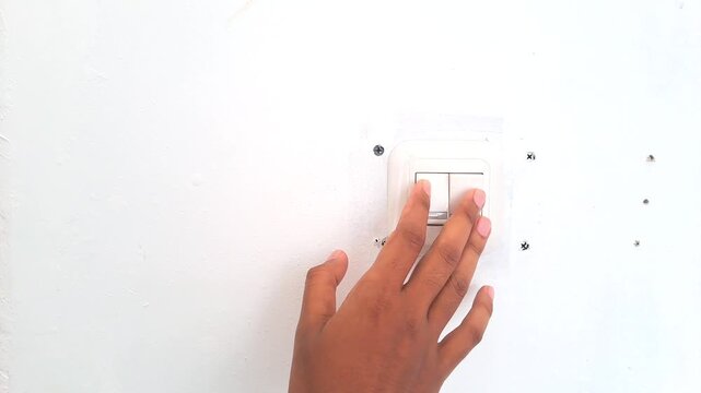 Close up of an Asian man hand turning on a white wall light switch in a minimalist workspace. Young male finger pressing the button to switch on the lights, initiating work or saving energy concept. 