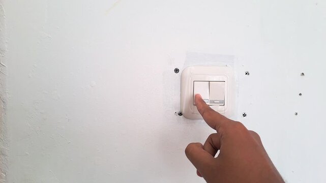 Close up of an Asian man hand turning on a white wall light switch in a minimalist workspace. Young male finger pressing the button to switch on the lights, initiating work or saving energy concept. 