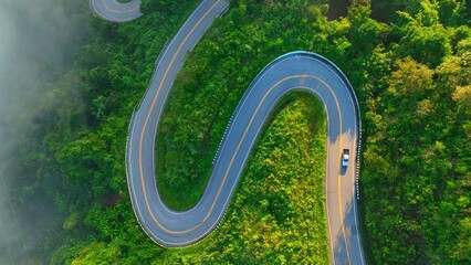 Obraz premium Aerial drone view of a stunning winding mountain road through a misty tropical forest at sunrise, with a car climbing the steep path, symbolizing transportation, and adventure. Nan Province, Thailand.