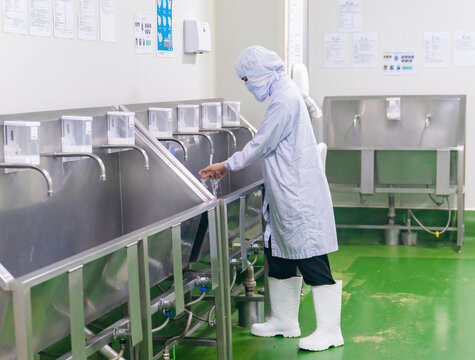 A food processing plant worker in full protective uniform, hairnet, mask, and white rubber boots washes their hands at a stainless steel industrial sink station before entering the production line.