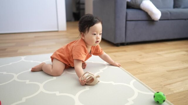 Soft light fills cozy living room scene. Baby crawls on patterned rug with green ball nearby. Child wears orange outfit and explores floor playfully. Gentle focus captures baby's curious
