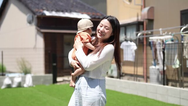 Sunlight warms a tidy garden yard. Woman holds baby, both smiling warmly. Baby looks up, curious and content. Joyful moment, perfect for family scenes. Homey, peaceful, natural setting