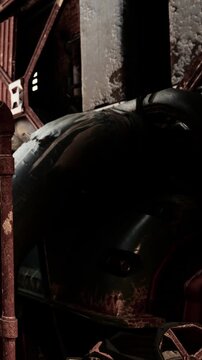 A large old vehicle sits in a warehouse. The place is dark and shadows cover most of the space. There is some equipment around, adding to the industrial look of the scene.