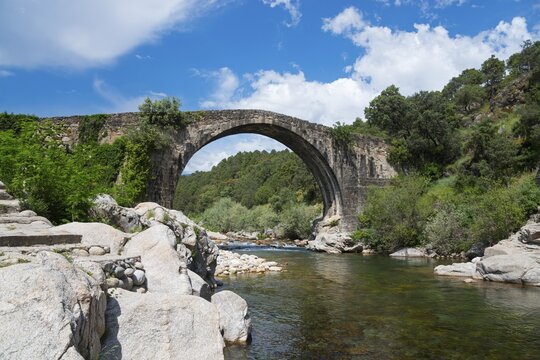 Steinbr&uuml;cke &uuml;ber einem ruhigen Fluss mit felsiger Uferlandschaft und dichtem Gr&uuml;n unter einem blau bew&ouml;lkten Himmel, R&ouml;mische Br&uuml;cke, Piscina natural de Madrigal de la Vera, Naturbad Madrigal de la Vera, Garganta de Alardos, Schlucht Alardos, C&aacute;ceres, Extremadura, Spanien