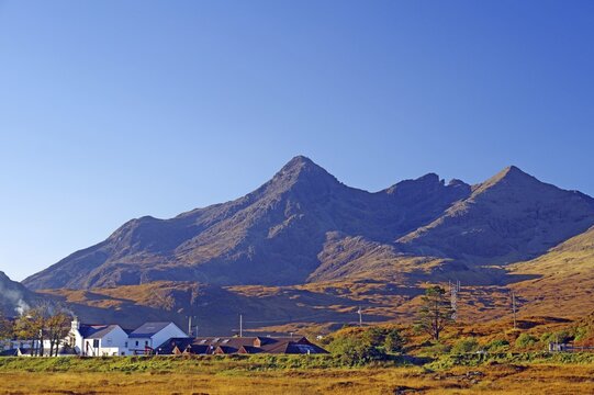A small village lies against an impressive mountain backdrop under a clear blue sky, October, Cullins, Hebrides, Isle of Skye, Hebrides, Scotland, United Kingdom