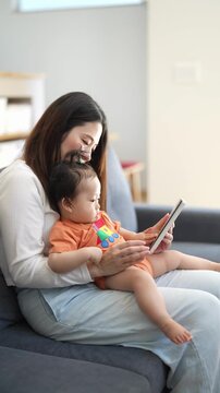 Mother smiles warmly while holding child. Child gazes at tablet with quiet curiosity. They sit close on soft couch, sharing quiet moment. Perfect for parenting, family, or tech use