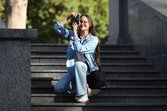 Young woman photographer taking pictures with vintage camera on city stairs outdoors, smiling female tourist in casual denim clothes capturing urban travel moments in sunny light