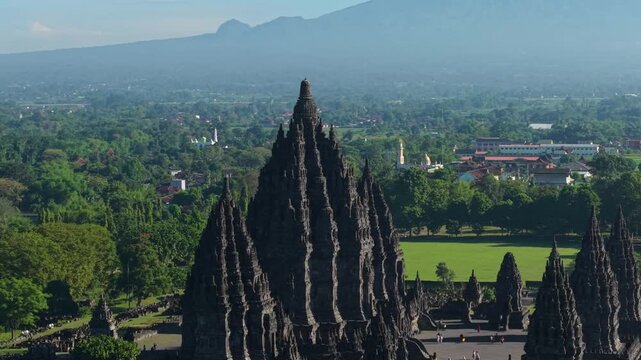 Aerial view of the ancient Prambanan Temple with towering stone spires surrounded by lush green lawns and a hazy mountain in Kecamatan Prambanan, Daerah Istimewa Yogya, Indonesia.