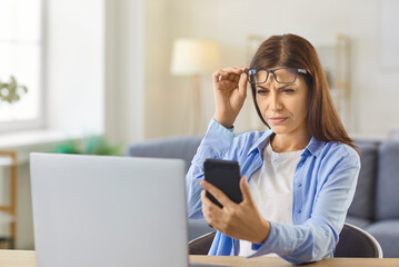 Woman with myopia, presbyopia and vision loss holding glasses on forehead to peer at and read text on mobile phone screen, struggle with blur and eyestrain. Person working at laptop at home desk © Studio Romantic